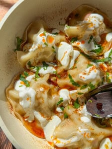 A close-up of Viral Blanket Dumplings topped with dollops of yogurt, a drizzle of red chili oil, chopped fresh herbs, and a sprinkle of spices, served in a bowl with a wooden spoon.