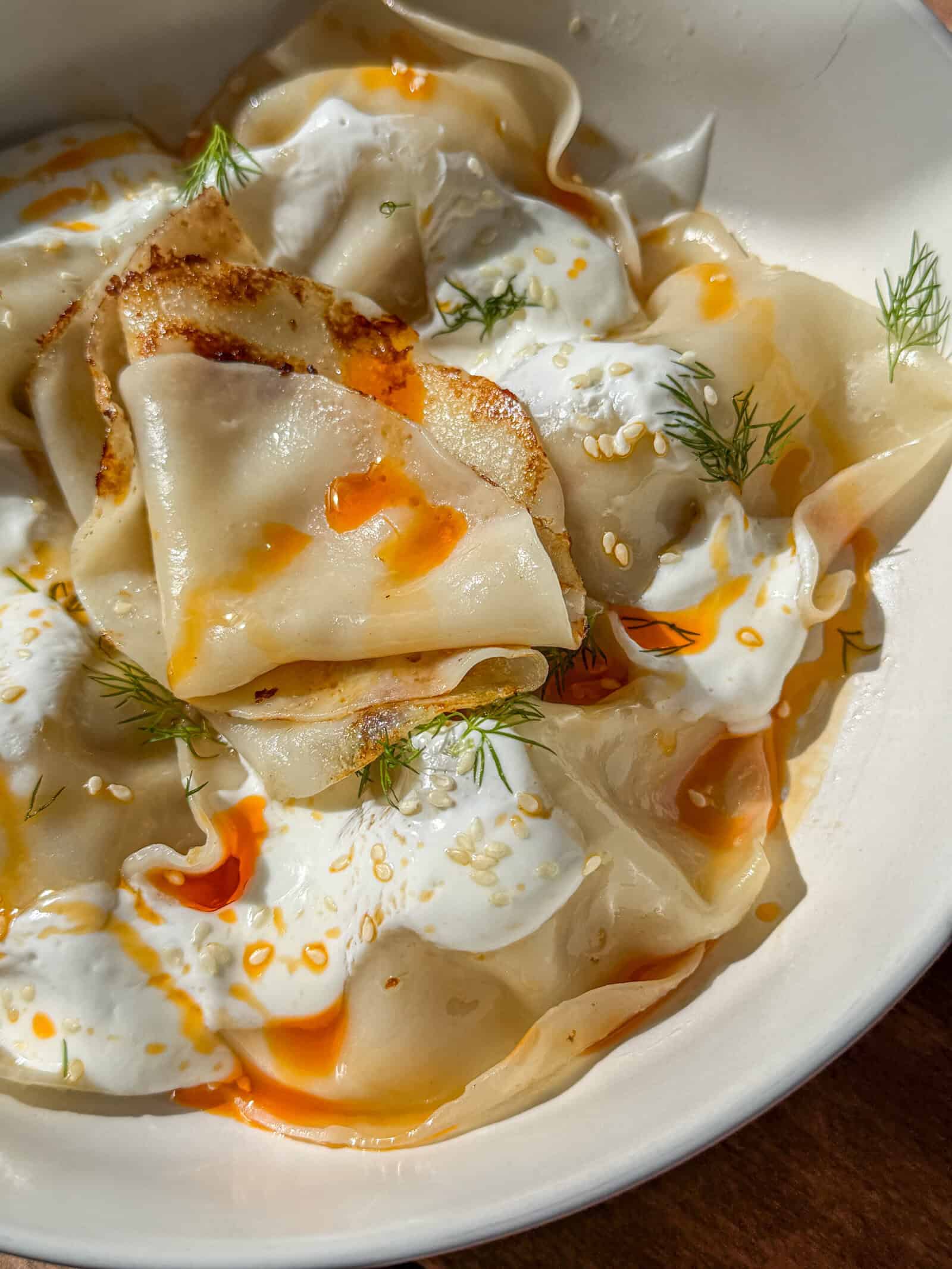 A close-up of Viral Blanket Dumplings topped with creamy white sauce, orange chili oil, sesame seeds, and fresh dill, served in a white bowl.