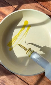 A stream of olive oil is being poured from a bottle into a light-colored frying pan, ready to cook the viral blanket dumplings, while sunlight casts shadows on the wooden surface.
