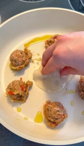A hand places a round dumpling wrapper over a scoop of meat and vegetable filling in a white pan with other filling scoops and drizzled oil, prepping Viral Blanket Dumplings to cook.