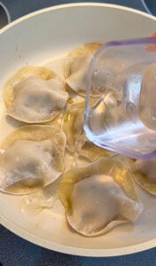 A hand pours water from a measuring cup into a skillet filled with Viral Blanket Dumplings, steaming or cooking these trendy potstickers to perfection.