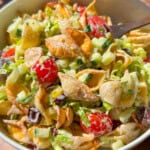 A bowl of pasta salad with shell pasta, cherry tomatoes, chopped cucumbers, olives, lettuce, and a creamy dressing, with a wooden spoon resting in the bowl. Sunlight brightens the colorful ingredients.