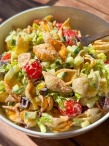 A bowl of pasta salad with shell pasta, cherry tomatoes, chopped cucumbers, olives, lettuce, and a creamy dressing, with a wooden spoon resting in the bowl. Sunlight brightens the colorful ingredients.