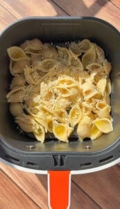 Uncooked pasta shells topped with shredded cheese and breadcrumbs in an air fryer basket, placed on a wooden surface with sunlight casting shadows.