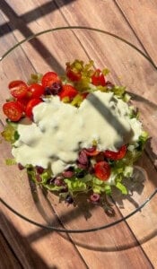 A glass bowl filled with chopped romaine lettuce, cherry tomatoes, sliced black olives, diced green peppers, and topped with a generous amount of creamy salad dressing, sitting on a wooden surface in sunlight.