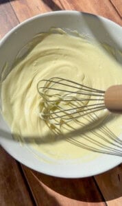 A white bowl filled with creamy yellow batter is being mixed with a metal whisk with a wooden handle, placed on a wooden surface in bright sunlight.