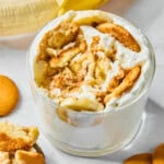 A glass filled with banana pudding topped with whipped cream, vanilla wafers, and banana slices. A spoonful of pudding rests nearby, along with whole vanilla wafers and a partially peeled banana in the background.