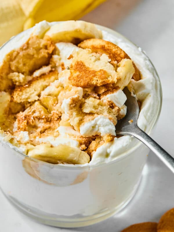 A close-up of a glass filled with banana pudding, whipped cream, banana slices, and crumbled vanilla wafers, with a spoon scooping out a portion. A banana and wafers are visible in the background.