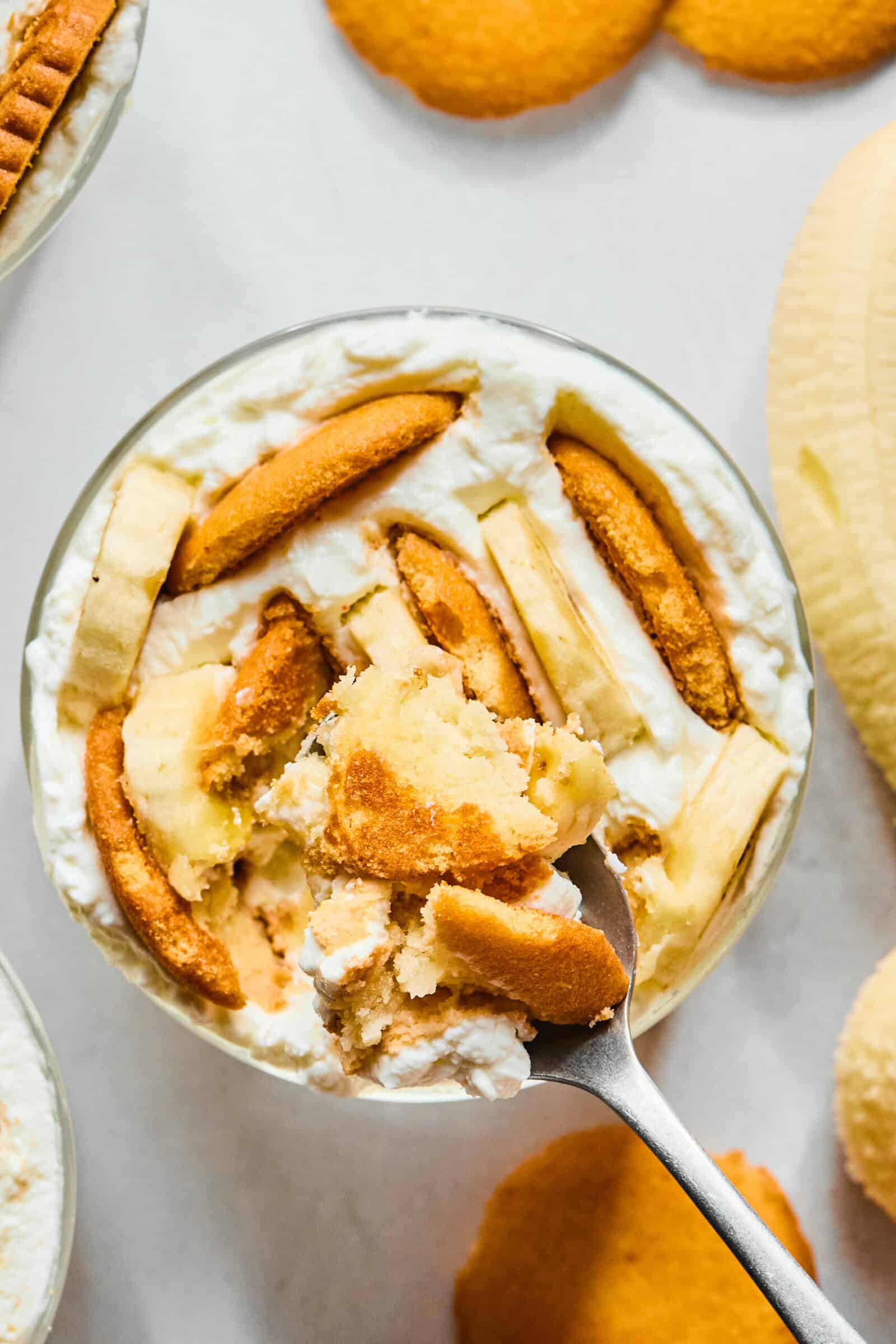 A close-up of banana pudding in a glass dish, topped with vanilla wafers and banana slices, with a spoon scooping out a portion. Vanilla wafers and a banana are visible in the background.