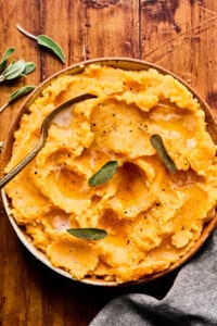 A bowl of creamy mashed sweet potatoes topped with melted butter, black pepper, and fresh sage leaves, set on a rustic wooden table with a metal spoon and a few sage sprigs nearby.