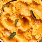 A bowl of creamy mashed potatoes topped with melted butter, cracked black pepper, and fresh sage leaves, with a spoon resting inside. The dish is set on a wooden surface.