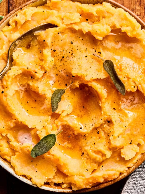 A bowl of creamy mashed potatoes topped with melted butter, cracked black pepper, and fresh sage leaves, with a spoon resting inside. The dish is set on a wooden surface.