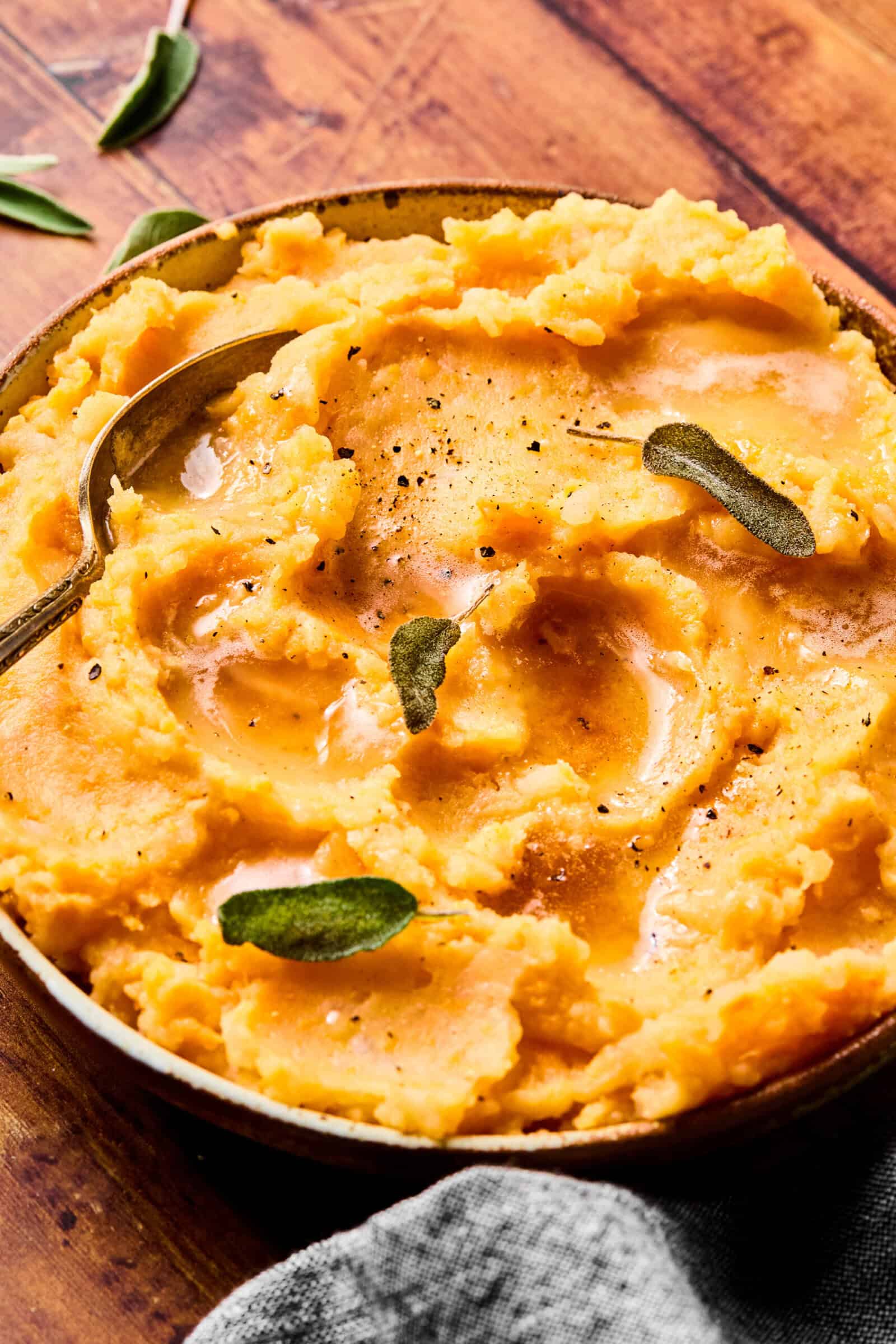 A bowl of creamy mashed sweet potatoes topped with gravy, black pepper, and fresh sage leaves, with a spoon resting in the dish, on a wooden surface.