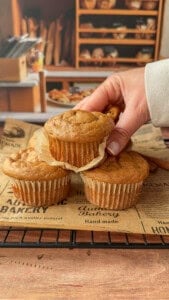 A hand lifts one of three muffins from a cooling rack lined with bakery-themed paper, with shelves of bread and baked goods visible in the background.