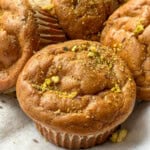 A close-up of golden-brown muffins topped with crushed pistachios, resting on a light-colored cloth. The muffins appear fluffy and moist, with slightly cracked tops and paper liners.