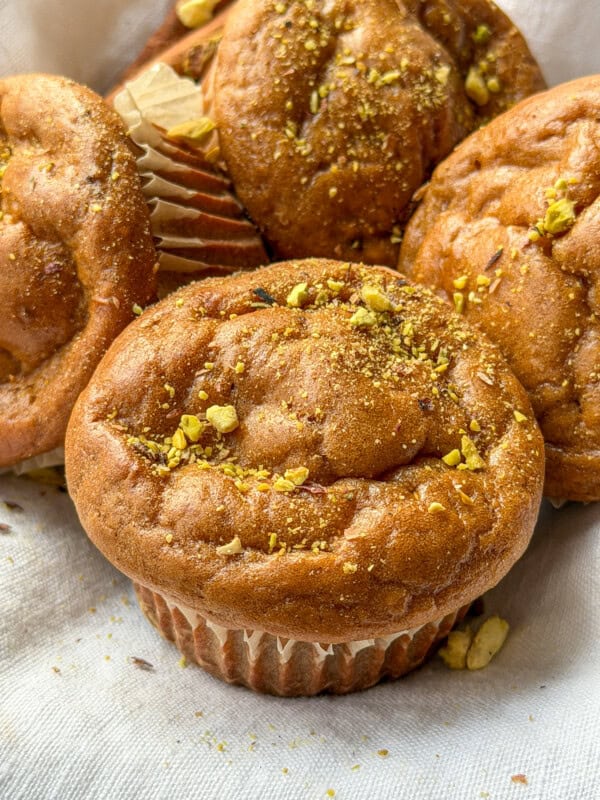 A close-up of golden-brown muffins topped with crushed pistachios, resting on a light-colored cloth. The muffins appear fluffy and moist, with slightly cracked tops and paper liners.