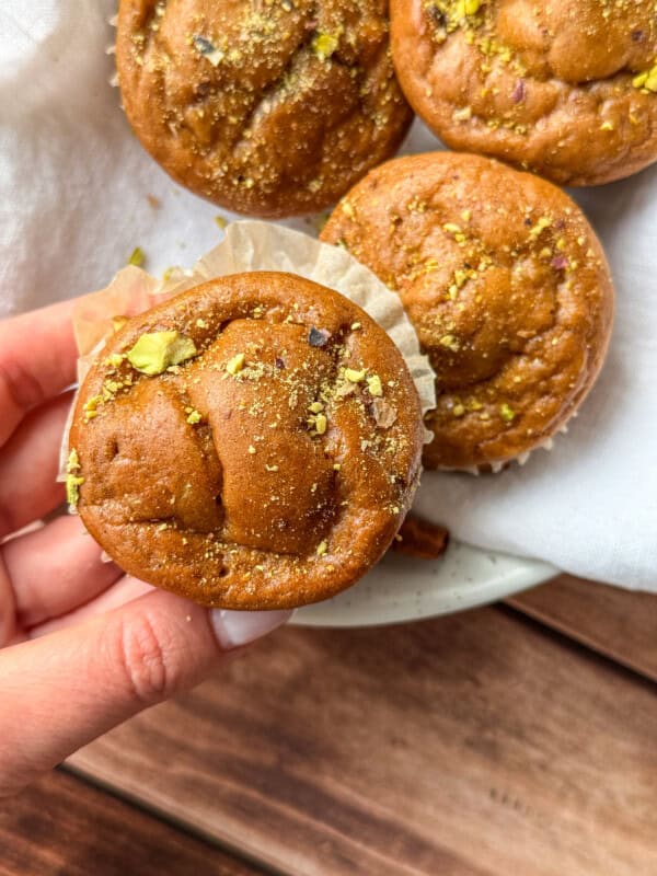 A hand holds a golden-brown muffin topped with crushed pistachios, with three more muffins and part of a white cloth visible in the background on a wooden surface.
