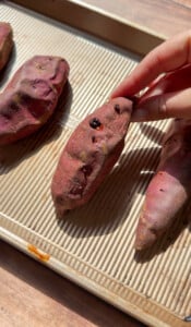 A hand holds a roasted sweet potato on a baking tray with other sweet potatoes, bathed in sunlight—perfect for making Healthy Sweet Potato Muffins.