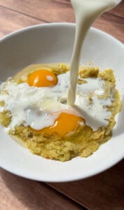 Milk being poured into a white bowl containing two raw eggs and mashed bananas, perfect for preparing Healthy Sweet Potato Muffins, set on a wooden surface.