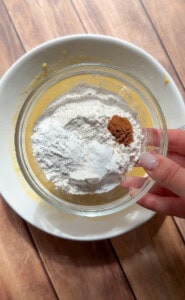 A hand holds a glass bowl containing flour, baking powder, and cinnamon above a larger white bowl with a yellow mixture on a wooden surface—perfect for making Healthy Sweet Potato Muffins.