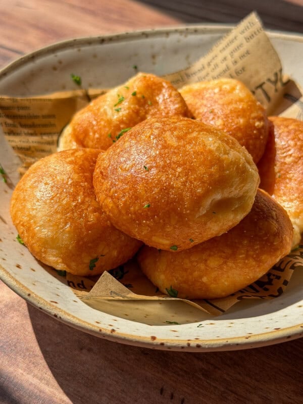 A ceramic bowl lined with parchment paper holds several golden-brown Air Fryer Parmesan Cheese Balls, sprinkled with herbs, and bathed in warm sunlight.