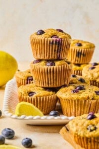 A stack of blueberry muffins in brown paper liners sits on a white plate, surrounded by fresh blueberries and lemon wedges—perfect alongside a Mediterranean Breakfast Wrap. More muffins and a whole lemon are visible in the background.