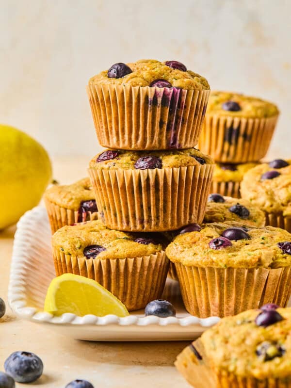 A stack of blueberry muffins in brown paper liners sits on a white plate, surrounded by fresh blueberries and lemon wedges—perfect alongside a Mediterranean Breakfast Wrap. More muffins and a whole lemon are visible in the background.