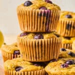 A stack of three blueberry muffins sits on a wooden surface, with several more muffins and a lemon wedge in the background—perfect alongside a Mediterranean Breakfast Wrap for a delightful and wholesome morning spread.