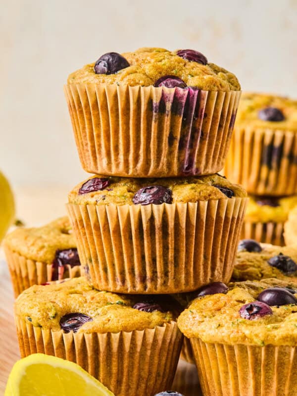 A stack of three blueberry muffins sits on a wooden surface, with several more muffins and a lemon wedge in the background—perfect alongside a Mediterranean Breakfast Wrap for a delightful and wholesome morning spread.
