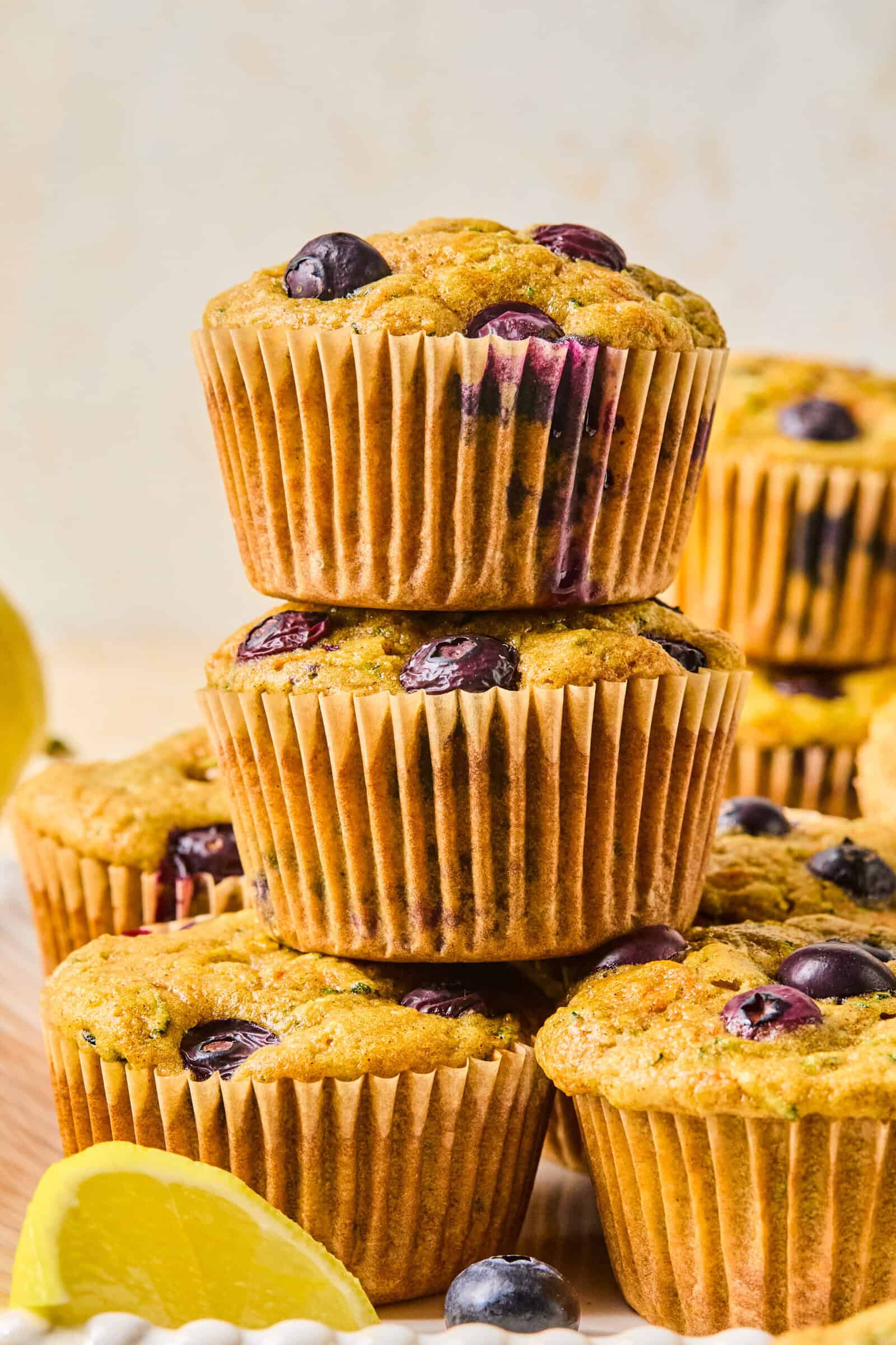 A stack of three blueberry muffins sits on a wooden surface, with several more muffins and a lemon wedge in the background—perfect alongside a Mediterranean Breakfast Wrap for a delightful and wholesome morning spread.