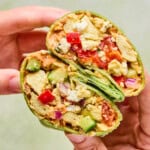 A hand holds a halved Mediterranean Breakfast Wrap filled with scrambled tofu, diced red and green bell peppers, onions, avocado, and other vegetables in a green spinach tortilla.