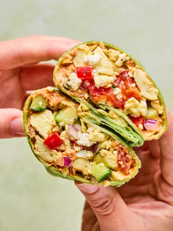 A hand holds a halved Mediterranean Breakfast Wrap filled with scrambled tofu, diced red and green bell peppers, onions, avocado, and other vegetables in a green spinach tortilla.