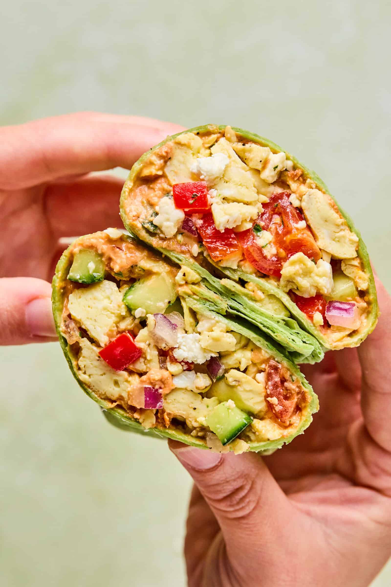 A hand holds a halved Mediterranean Breakfast Wrap filled with scrambled tofu, diced red and green bell peppers, onions, avocado, and other vegetables in a green spinach tortilla.