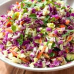 A bowl of colorful coleslaw featuring chopped green and purple cabbage, shredded carrots, and fresh herbs, with a wooden spoon resting in the bowl on a wooden table.