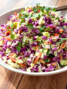 A bowl of colorful coleslaw featuring chopped green and purple cabbage, shredded carrots, and fresh herbs, with a wooden spoon resting in the bowl on a wooden table.