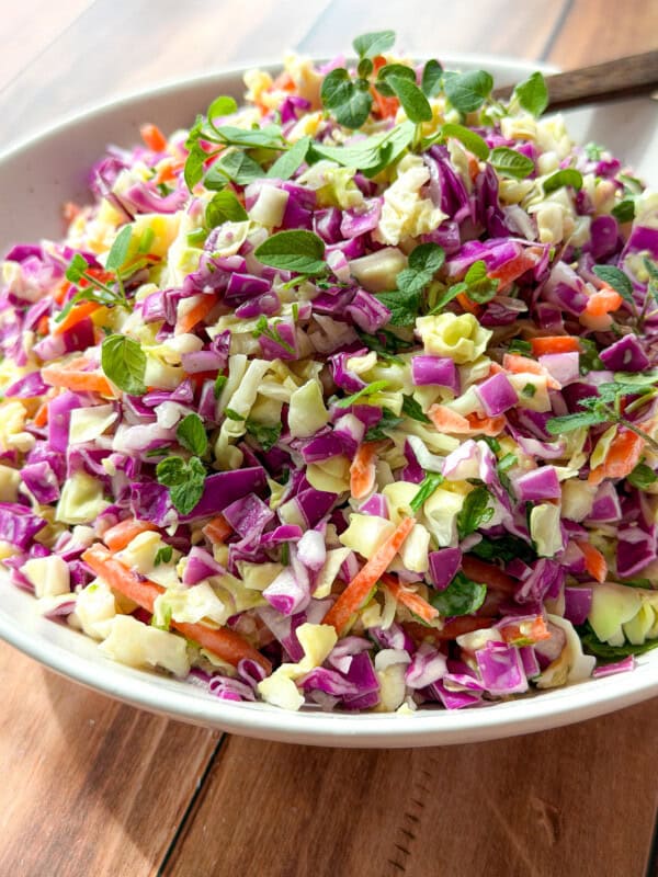 A bowl of colorful coleslaw featuring chopped green and purple cabbage, shredded carrots, and fresh herbs, with a wooden spoon resting in the bowl on a wooden table.
