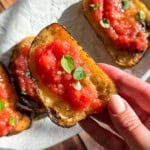 A hand holds a slice of toasted bread topped with crushed tomatoes and fresh green herbs, reminiscent of flavors from an Italian eggplant parmesan recipe, with more slices on a white plate in the background on a sunlit wooden surface.