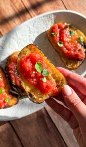 A hand holds a slice of toasted bread topped with crushed tomatoes and fresh green herbs, reminiscent of flavors from an Italian eggplant parmesan recipe, with more slices on a white plate in the background on a sunlit wooden surface.