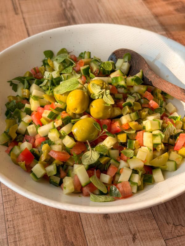 A white bowl filled with a chopped salad of cucumbers, tomatoes, green peppers, and green olives, garnished with fresh herbs. Perfect alongside a classic bread dipping oil recipe. Two wooden spoons rest on the side, atop a wooden table.
