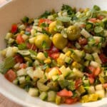 A bowl of chopped salad with cucumbers, tomatoes, green olives, pickles, and fresh herbs is served in a white dish on a wooden surface—perfect alongside your favorite bread dipping oil recipe.