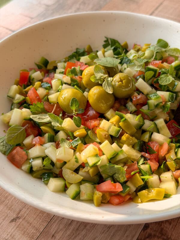 A bowl of chopped salad with cucumbers, tomatoes, green olives, pickles, and fresh herbs is served in a white dish on a wooden surface—perfect alongside your favorite bread dipping oil recipe.