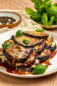 A plate of baked Eggplant Parmesan layered with melted cheese and tomato sauce, garnished with fresh basil leaves. A bunch of fresh basil and a dish of peppercorns sit in the background—perfect for trying your favorite eggplant parmesan recipe.