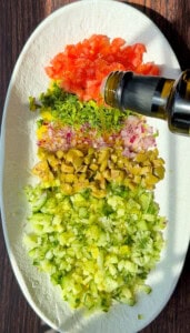 An overhead view of a plate with chopped tomatoes, herbs, onions, olives, and cucumbers arranged in rows, as olive oil is being poured over the top for a fresh bread dipping oil recipe.