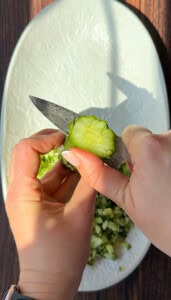 Close-up of hands holding a knife and slicing a cucumber over a white plate with chopped vegetables, perfect for pairing with your favorite bread dipping oil. Sunlight casts shadows on the plate and wooden surface beneath.