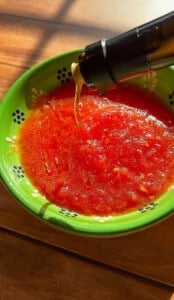 A stream of olive oil is being poured into a green bowl filled with grated tomatoes, ready for an olive oil bread dip. The bowl sits on a wooden surface and features a decorative pattern with small white flowers and blue dots.