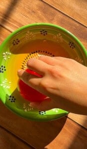 A hand holds a red tomato over a colorful green and yellow bowl with floral patterns, perfect for serving bread dipping oil, all set on a wooden surface in bright sunlight.