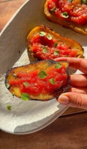 A hand holds a slice of toasted bread topped with tomato sauce and fresh herbs, perfect for pairing with your favorite olive oil bread dip. More slices with crispy, charred edges sit on a white plate in the background.