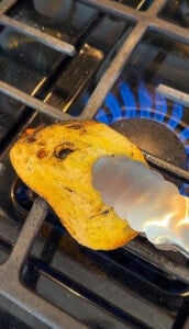 A piece of bread is held with metal tongs over an open gas stove flame, being toasted directly on the burner grate, perfect for enjoying with a homemade bread dipping oil recipe.