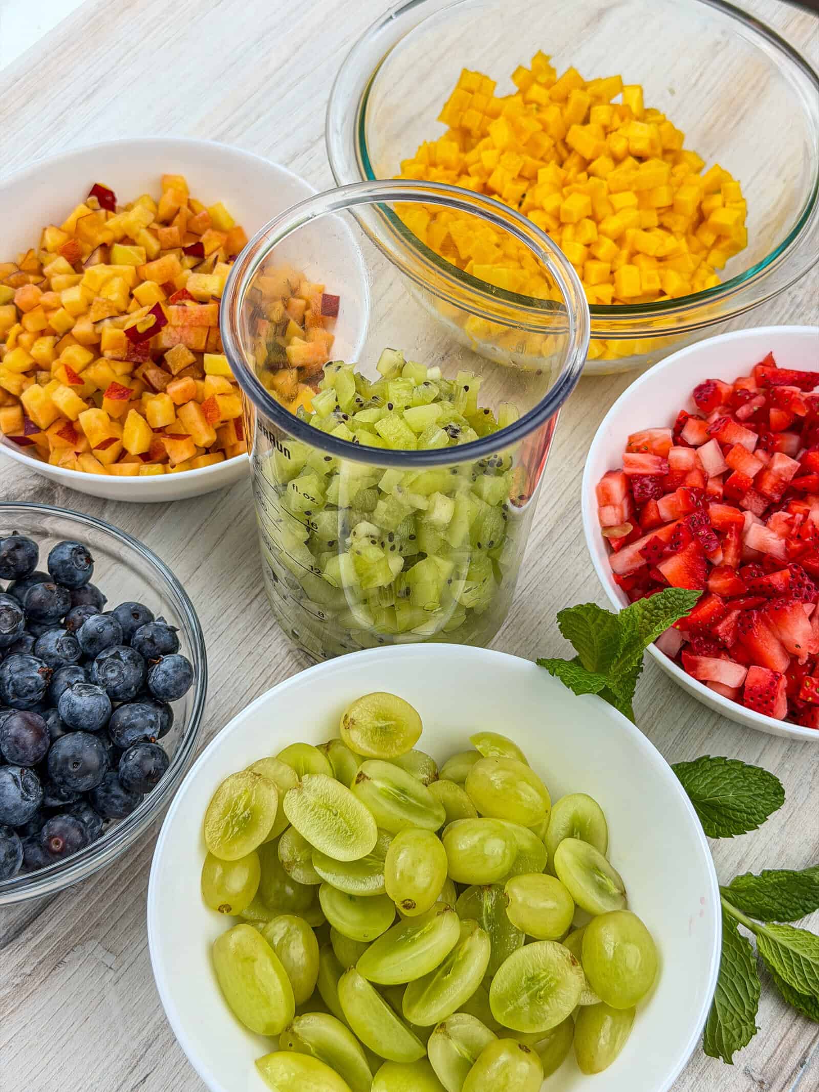 Bowls of chopped fruit on a light surface, including blueberries, green grapes, strawberries, kiwi, peach, and mango. There are also a couple of sprigs of fresh mint near the bowls.