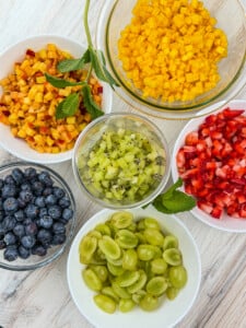 Six bowls of chopped fruit on a light wooden surface, including diced mango, diced peaches, chopped strawberries, blueberries, sliced green grapes, and diced kiwi, with mint leaves for garnish.